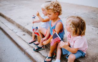 Three toddlers sit on the street curb with patriotic flags during a 4th of July celebration in the Texas Hill Country.