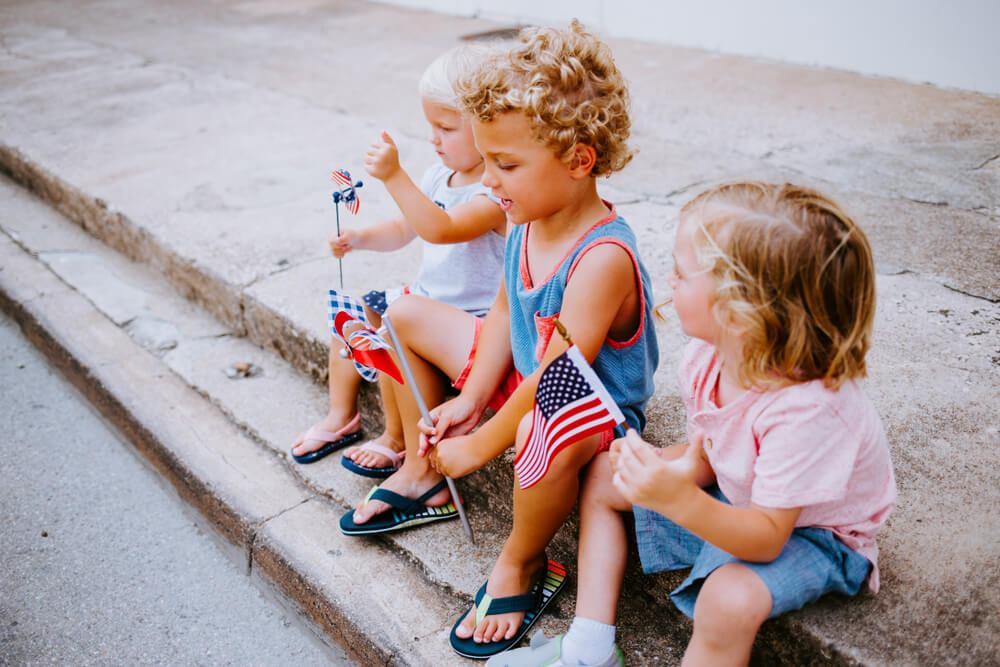 Three toddlers sit on the street curb with patriotic flags during a 4th of July celebration in the Texas Hill Country.